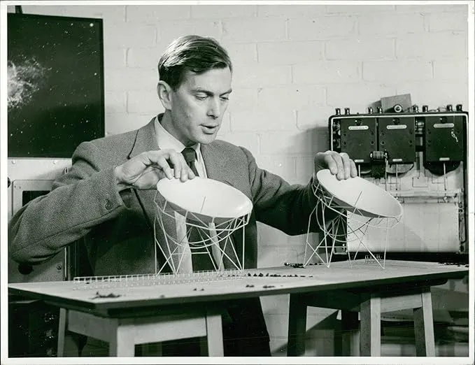 A black-and-white photograph shows a man in a suit jacket and tie leaning over a table, adjusting two small, white scale models of satellite dishes mounted on wire scaffolding. He looks down at the models with both hands resting lightly on the rims of the dishes. Behind him, a light-colored painted brick wall serves as the backdrop, flanked by a dark framed star map on the left and technical equipment with dials on the right.