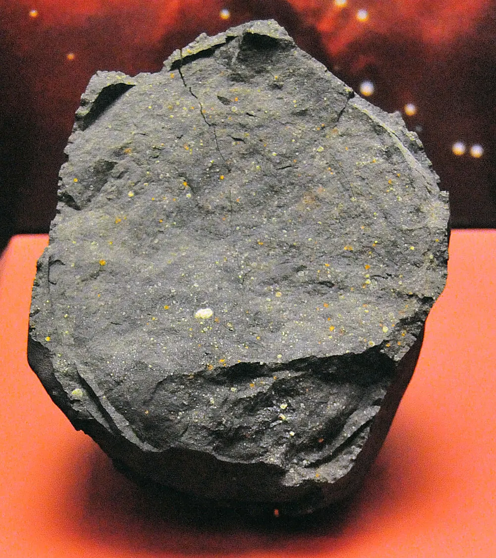 A large, dark charcoal-grey rock with a rough, cracked surface sits centrally on a bright orange platform. Tiny flecks of yellow and orange mineral specks are scattered across its flat, mat-like face, with a fine crack running vertically near the top. The background features a soft-focus, deep red hue dotted with small, glowing white blurred spots.