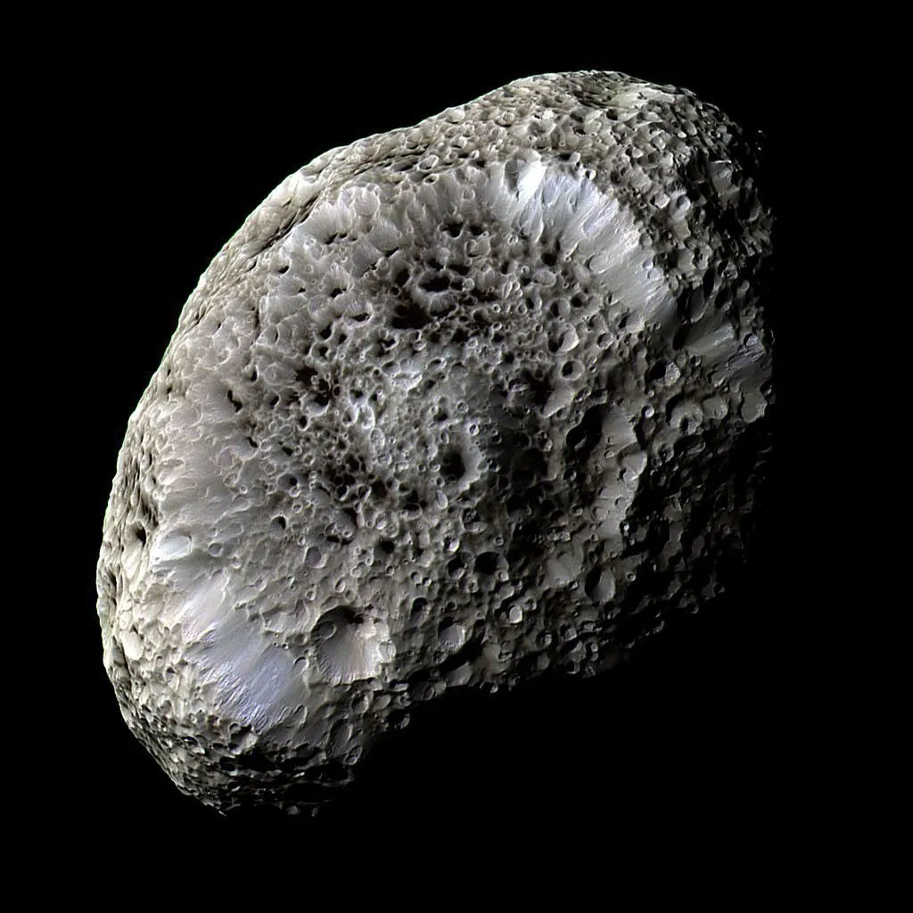An irregularly shaped, sponge-like celestial body with a heavily pitted, crater-covered surface fills the center of the frame against a solid black space background. The pale grey and light-tan object features deep, honeycomb-like depressions and several prominent larger circular basins with bright, sunlit rims. Strong directional lighting casts crisp dark shadows inside the craters, emphasizing its rough, porous texture and sharp contrast against space.
