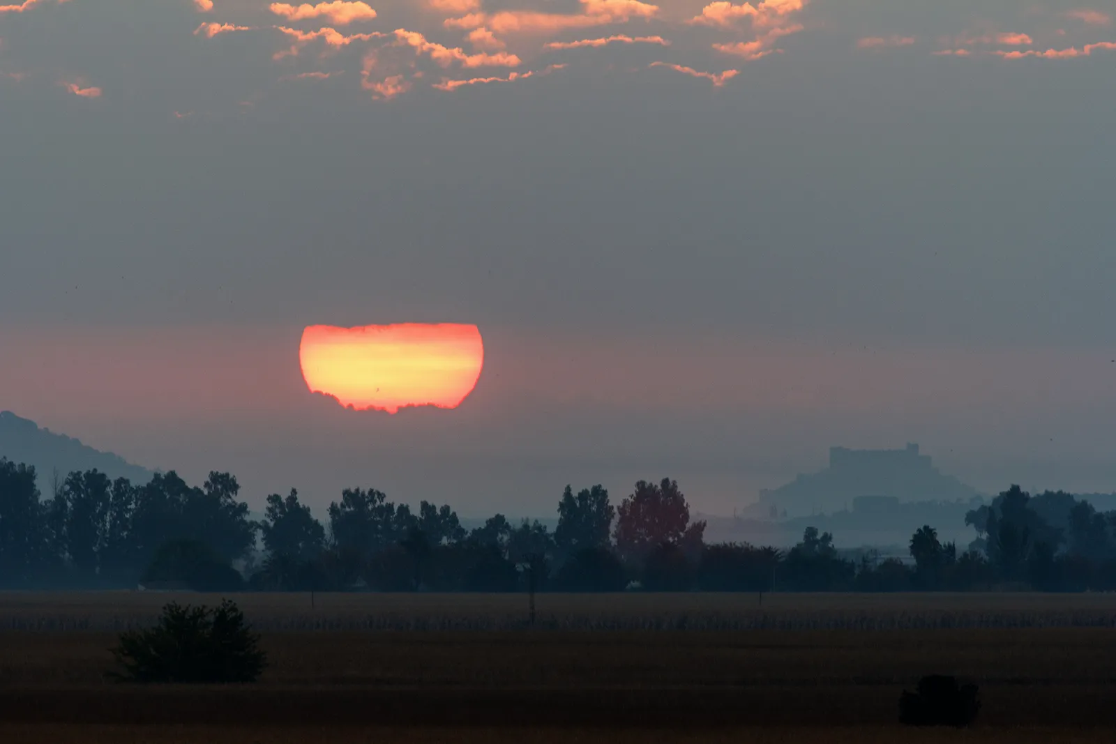 A photograph of the autumnal equinox sunrise over a hazy rural landscape in Spain, showing the Sun as a large, heavily distorted red-orange disc flattened by atmospheric refraction just above the treeline. A silhouetted tree line, distant mountains, and what appears to be a castle or fortress on a hilltop are visible through the haze. Pink-tinted clouds and a gray-blue sky fill the upper portion of the frame, and a flat golden field stretches across the foreground.