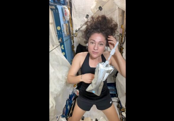 a woman wearing a black tank top and black shorts holds a water bottle on board a space station