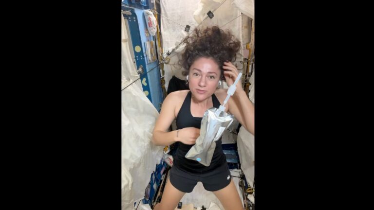a woman wearing a black tank top and black shorts holds a water bottle on board a space station