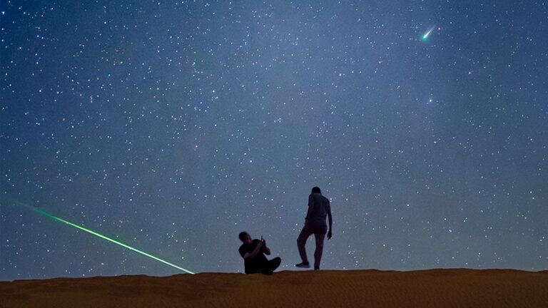 Two stargazers are silhouetted standing on a sand dune beneath a starry sky. A comet is visible to the upper right and a green meteorite can be seen streaking towards the ground to the left.