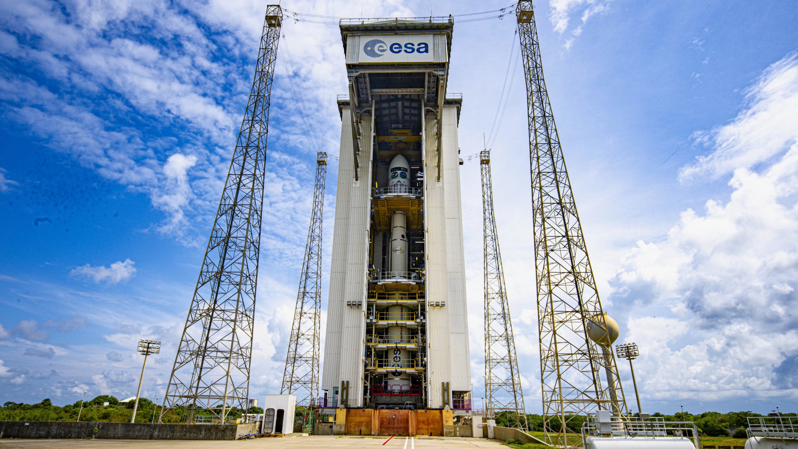 A small white rocket stands vertically inside a white assembly building under cloudy blue skies