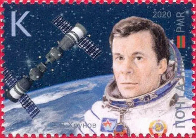 A rectangular postage stamp with a red perforated border features an illustrated portrait of a man in a white spacesuit filling the right foreground. To the left, two connected grey spacecraft with solar panels extend diagonally above a blue-and-white view of Earth against a dark starry sky. Bold lettering, numerals, and small colored flags frame the upper corners and right side under even, bright lighting.