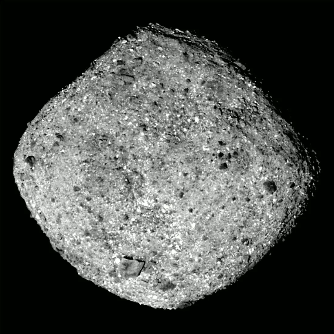 A monochrome, close-up photograph shows a diamond-shaped asteroid with a rough, gravel-like surface centered against a solid black background. Bright, direct lighting highlights thousands of tiny, reflective grey and white specks interspersed with dark, pitted shadows and scattered boulders. High contrast emphasizes its jagged, rounded perimeter and uneven, rubble-covered terrain.