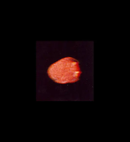 A small, blurry, strawberry-shaped blob glowing in warm orange and red tones sits centered within a dark square inset. Two faint, yellowish points of brightness stand out along its right edge, contrasting against the muted red body. The entire square frame is isolated against a solid black background, giving the object high contrast.