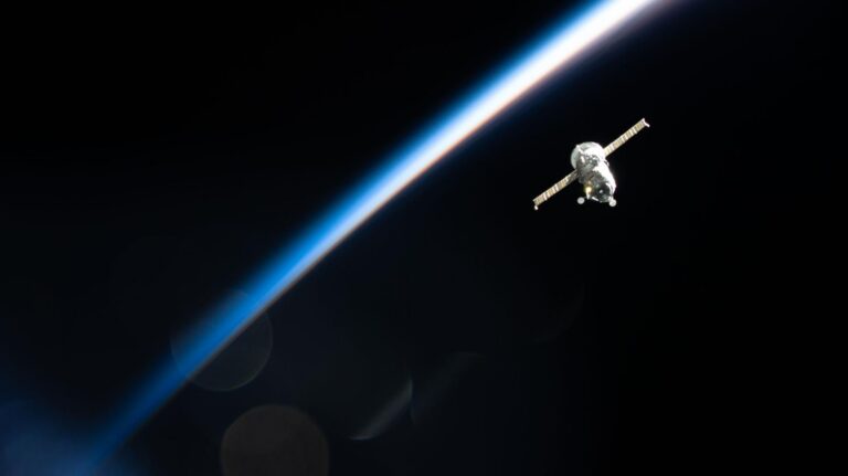 photo of a spacecraft orbiting a mostly dark earth, whose atmosphere is lit up and shows up as a bright bluish-white arc