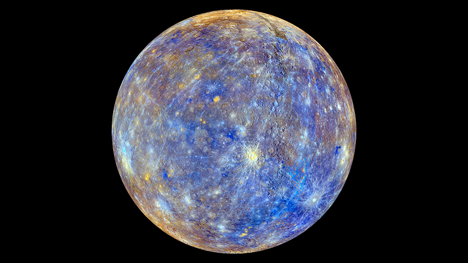 A fully illuminated sphere showing a planet-like body sits centered against a solid black background. Its surface is mottled with vibrant shades of deep blue, tan, gold, and light grey, covered in circular impact craters and ray-like streaks. A bright pale-blue crater near the upper-left center shoots thin, light-colored lines outward across the surrounding dark blue and tan terrain.