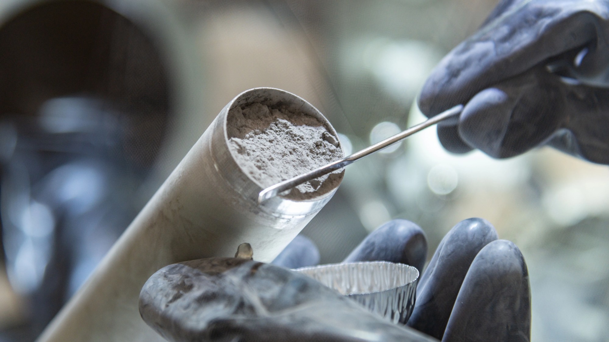 a person's hands in rubber gloves scrapes grey dust out of a metal tube into a foil tray