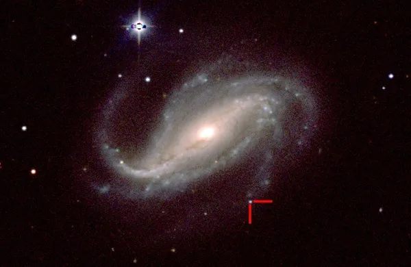 A broad, S-shaped spiral formation with a brightly glowing yellow-white core rests tilted at the center of a dark, starry sky. Two faint, hazy blue arms curve outward from the center, surrounded by smaller, scattered pinpricks of white light. Near the lower right curve of the outer arm, two small red lines form a right-angle marker pointing to a single bright white star-like dot.
