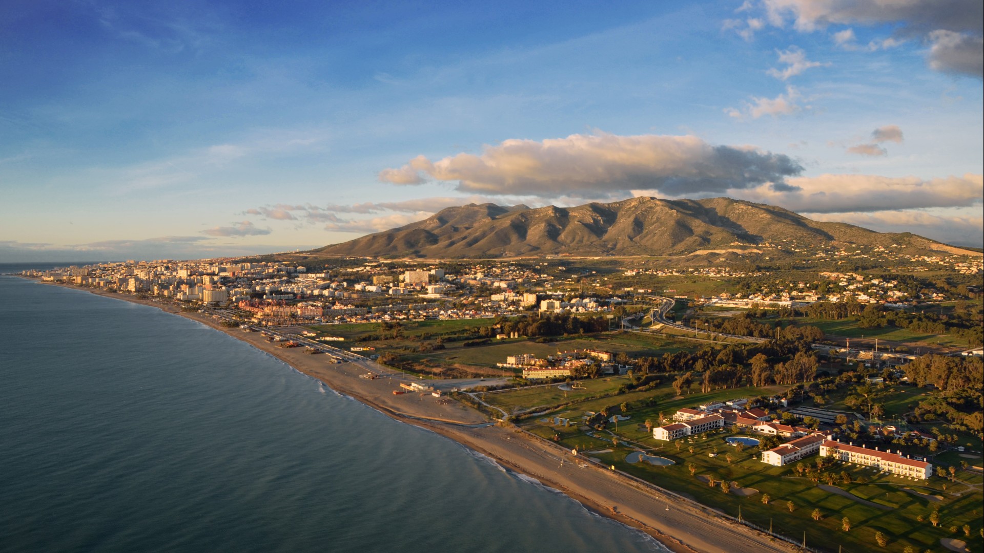 Aerial view of the Mediterranean coast. There are large hills in the background, looming over the city below. Above is a blue sky, dotted with large clouds.