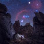 A night sky showing the constellation Orion peaking between large craggy rocks. At the bottom is a human holding up a light.