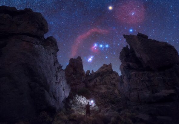 A night sky showing the constellation Orion peaking between large craggy rocks. At the bottom is a human holding up a light.