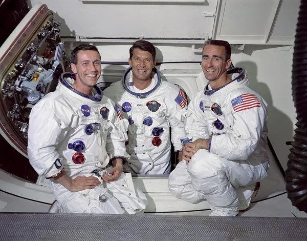 A color photograph shows three smiling men wearing white spacesuits adorned with patches and American flag emblems, posing closely together inside a white spacecraft interior. The suit details, including blue and red metallic connectors, stand out brightly under clear, even indoor lighting. An open hatch door with metallic mechanical components frames the left side of the background.