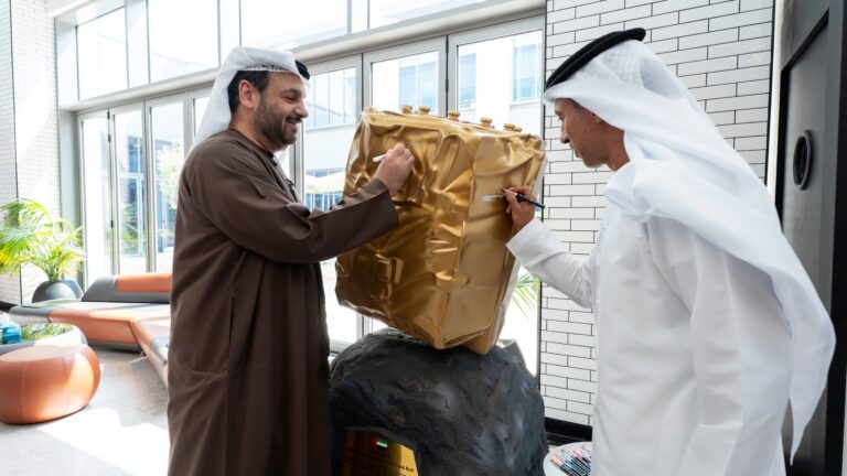 two men in long, flowing robes sign a model of a small golden spacecraft in a well-lit room