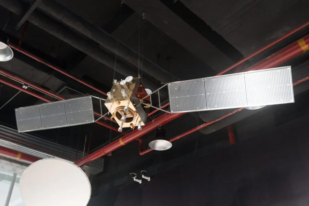 A metallic, box-shaped satellite model hangs suspended in the center against a dark industrial ceiling crossed with red pipes. Two large, grid-patterned rectangular solar panels extend outward from opposite sides like wings, while small circular dishes and thruster nozzles attach to its brassy body. Direct overhead lighting reflects off the metallic surfaces and the white, grid-textured solar panels, creating moderate contrast with the shadowy ceiling behind it.