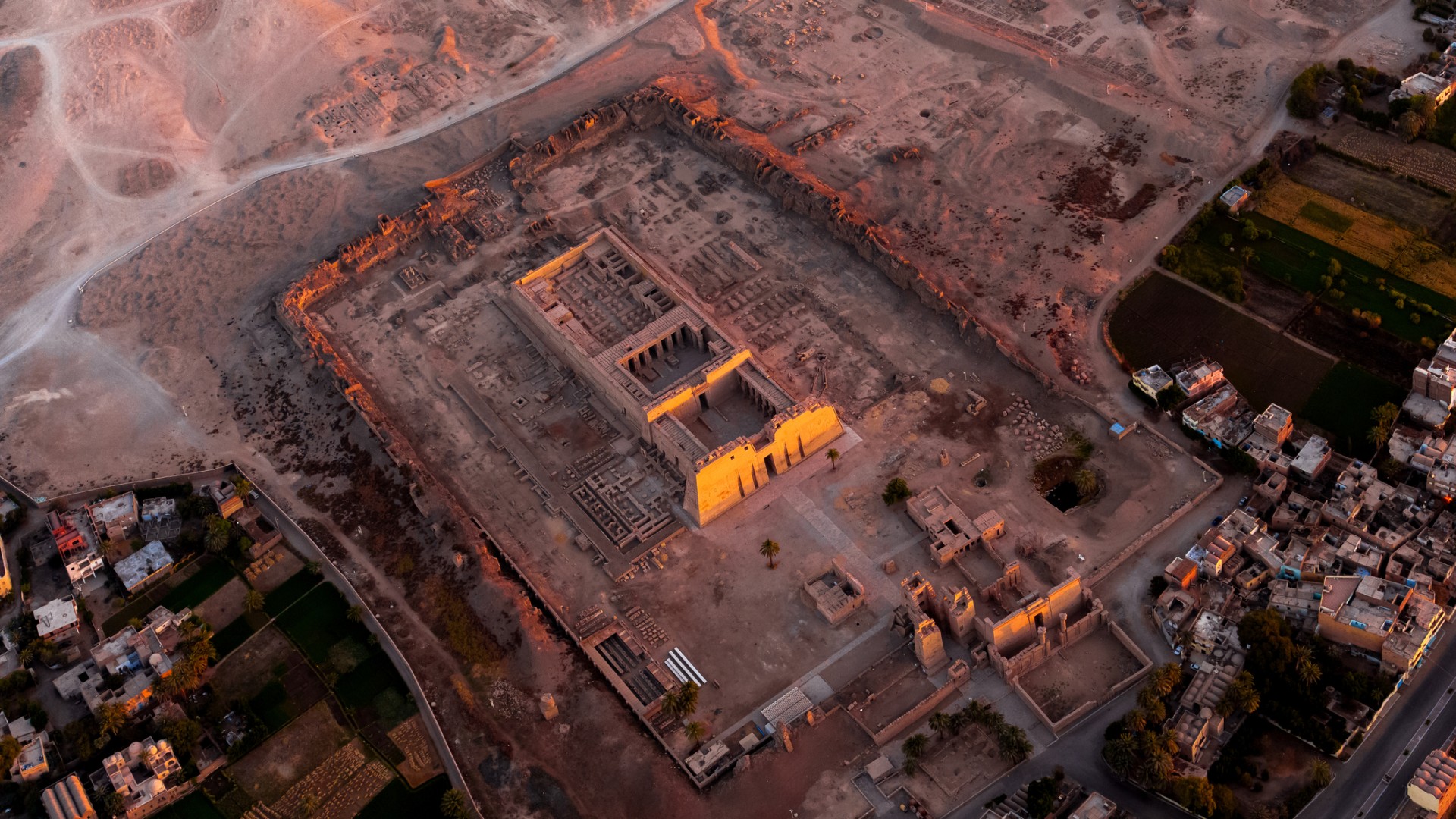 Aerial view of an ancient archaeological complex in a dusty landscape, with orange-lit stone ruins and nearby homes at dusk; atmospheric and solemn.