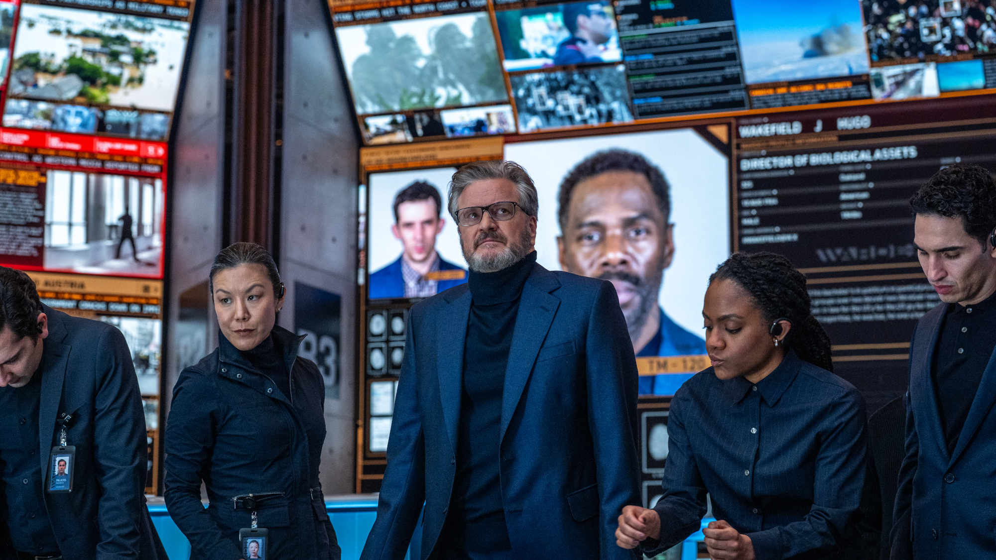 A group of agents in suits standing in front of security monitors.