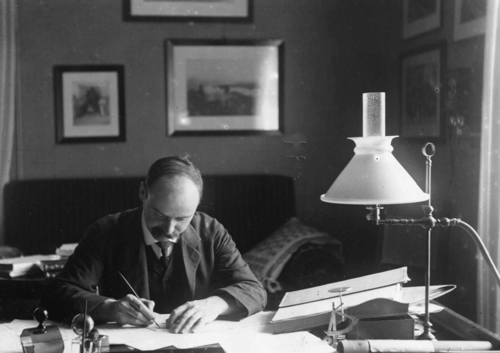 A black-and-white photograph shows a man in a dark suit seated at a desk, leaning forward as he writes on paper with a pen. A desk lamp with a light-colored, conical shade stands on the right, illuminating the documents and papers spread across the desktop. Framed pictures hang on the dark wall behind him, creating a focused, warm lighting contrast on the workspace.