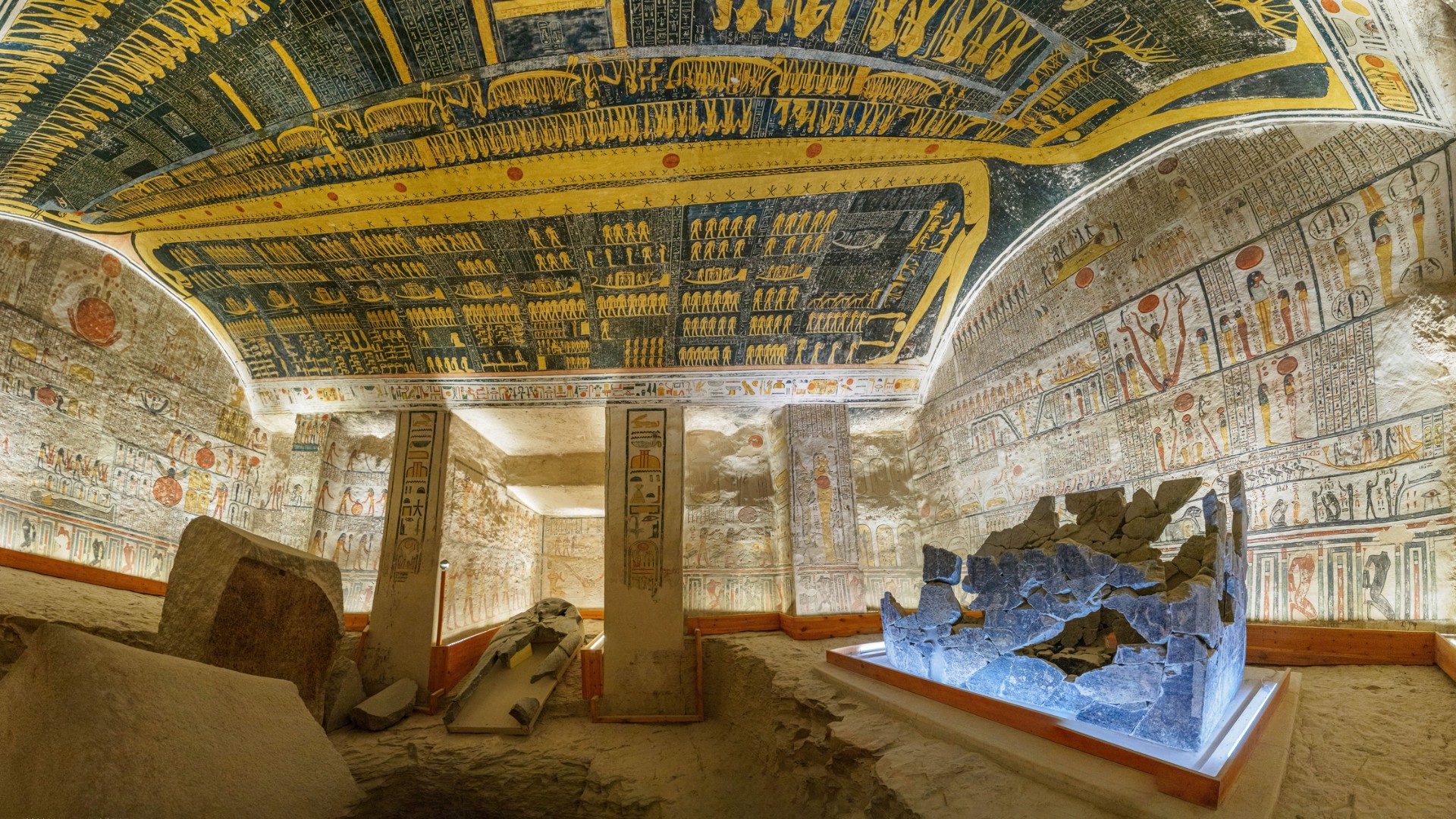 Interior of an ancient Egyptian tomb with colorful hieroglyphs and astronomical paintings on the ceiling and walls, featuring stone pillars and fragments of a sarcophagus.