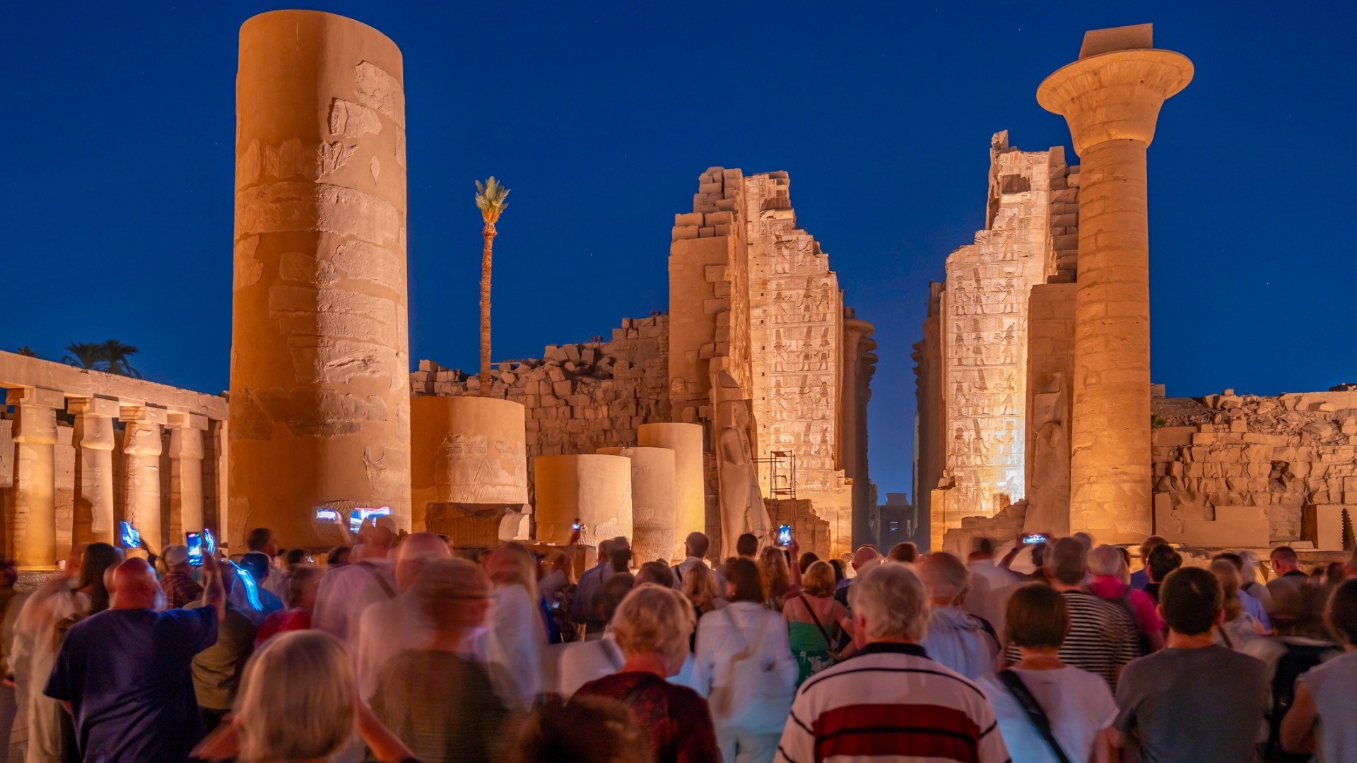 A group of people are gathered in front of the tall columns of the Karnak Temple at dusk.