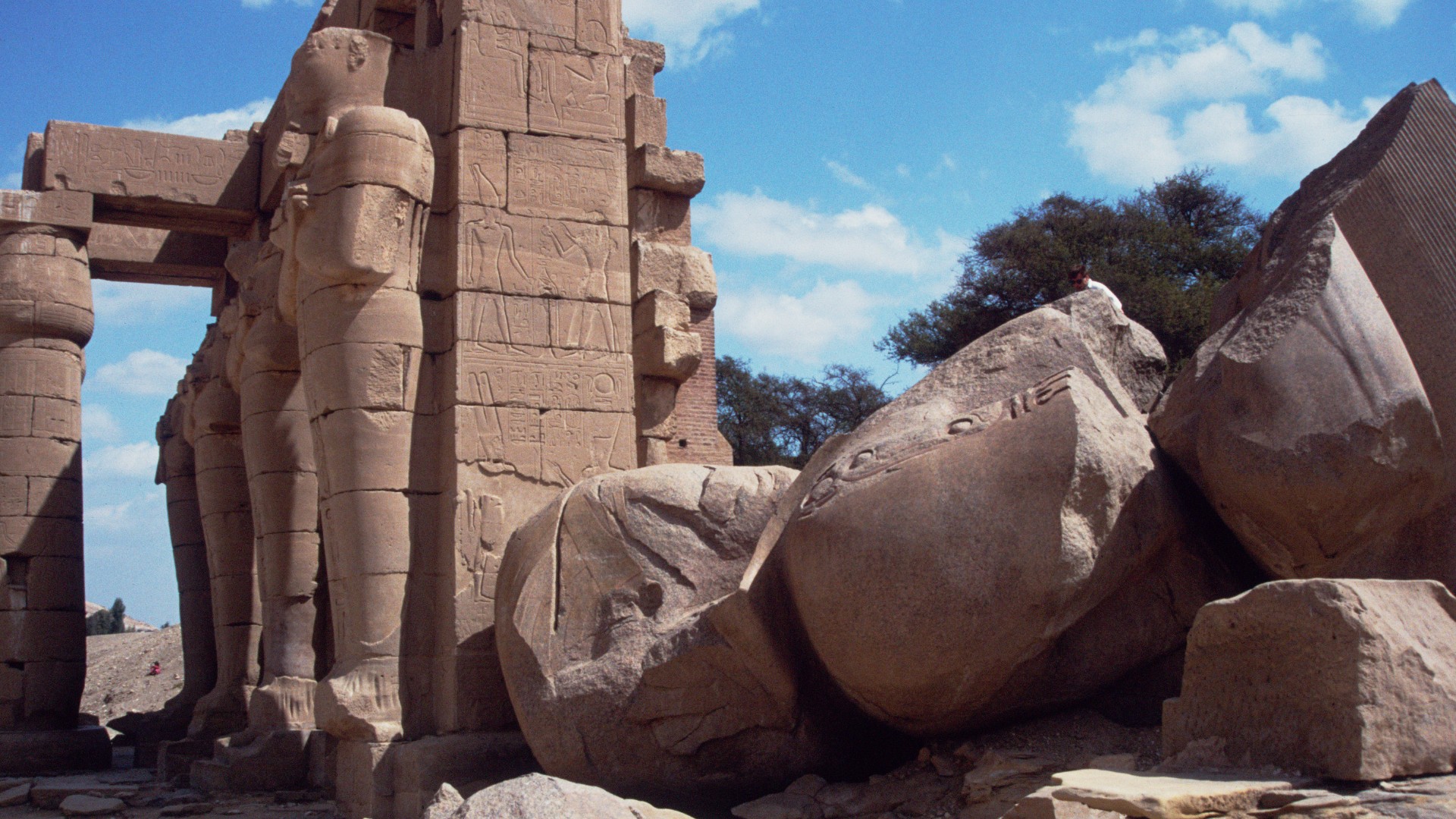 Sunlit ancient Egyptian temple ruins, with towering carved columns and human statues. AT the forefront is a giant fallen statue broken to pieces.