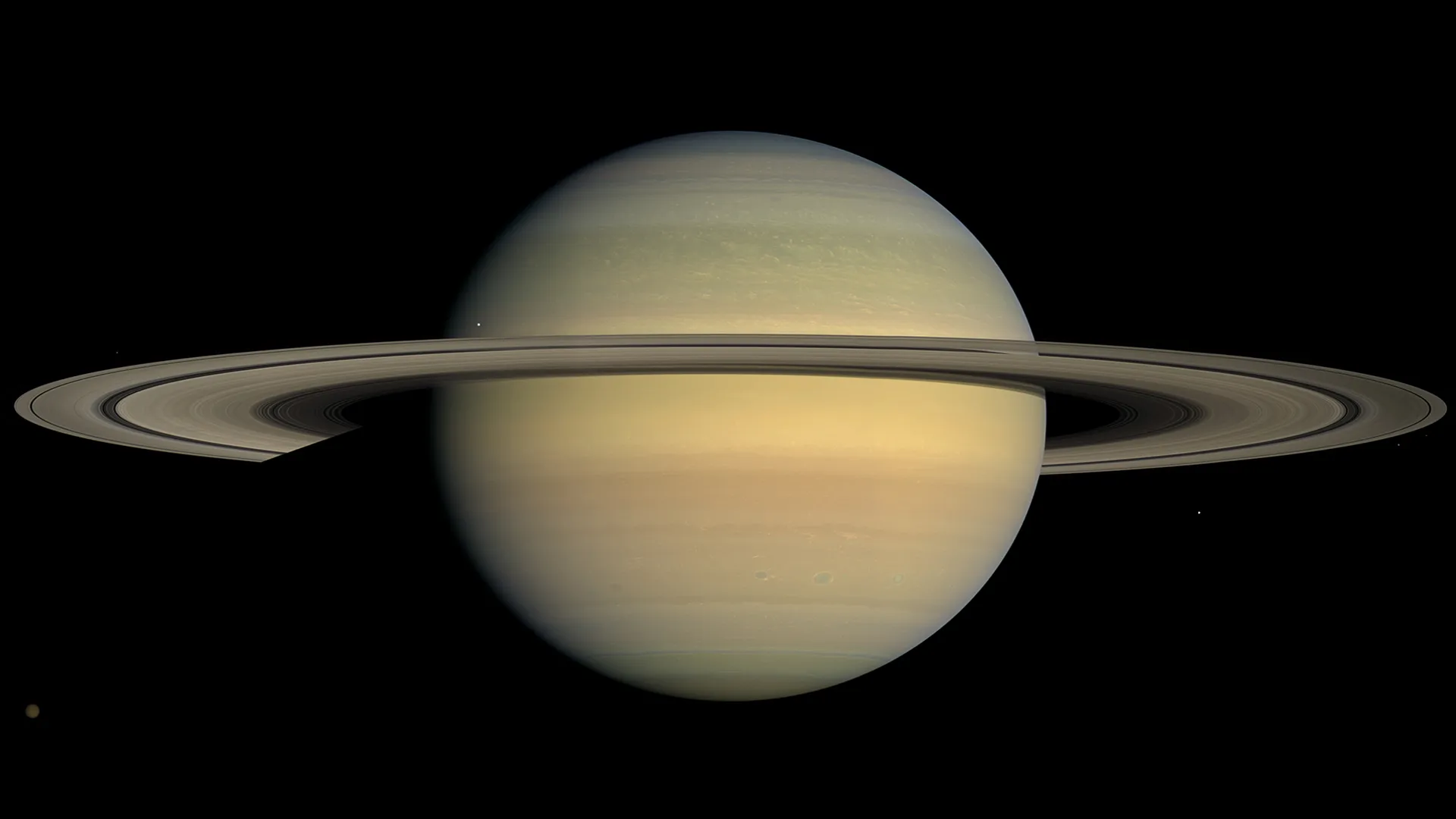 Photograph of Saturn against a black background, shown nearly face-on with pale cream and tan cloud bands crossing its globe and a faint orange tint near the equator. The wide ring system extends horizontally across the frame, and several small moons appear as faint points of light scattered around the planet.