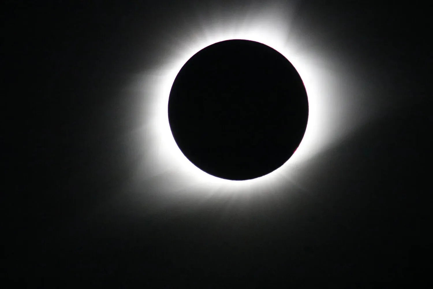 A stark, high-contrast photograph captures a solid black circle perfectly centered against a jet-black background. A bright, glowing ring of white light radiates outward from behind the dark circle, creating a soft, hazy halo with faint streaks extending into the surrounding darkness.