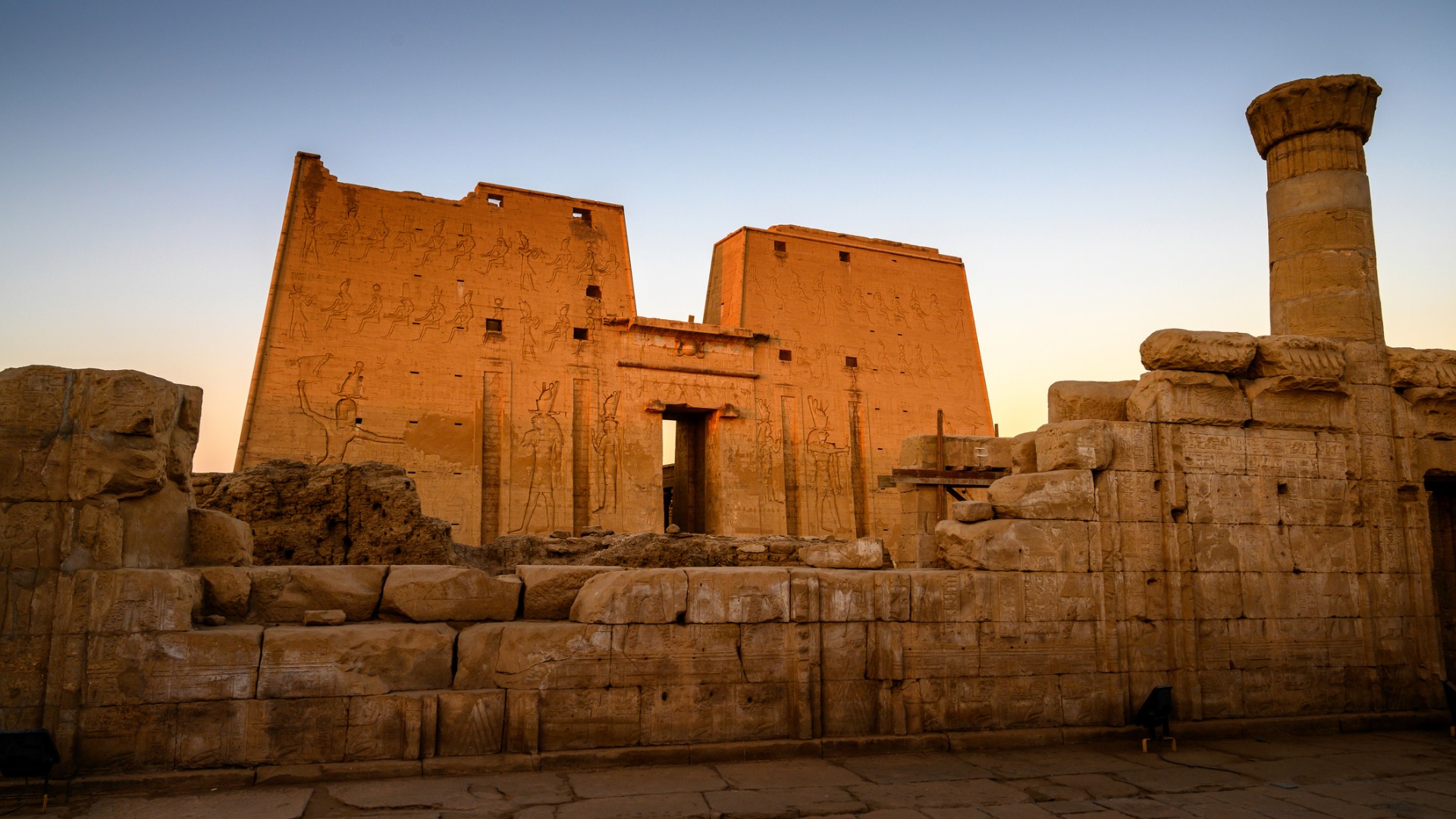 Ancient Egyptian temple ruins glow in warm sunset light, with carved walls, hieroglyphs, scattered stone blocks, and a tall column.