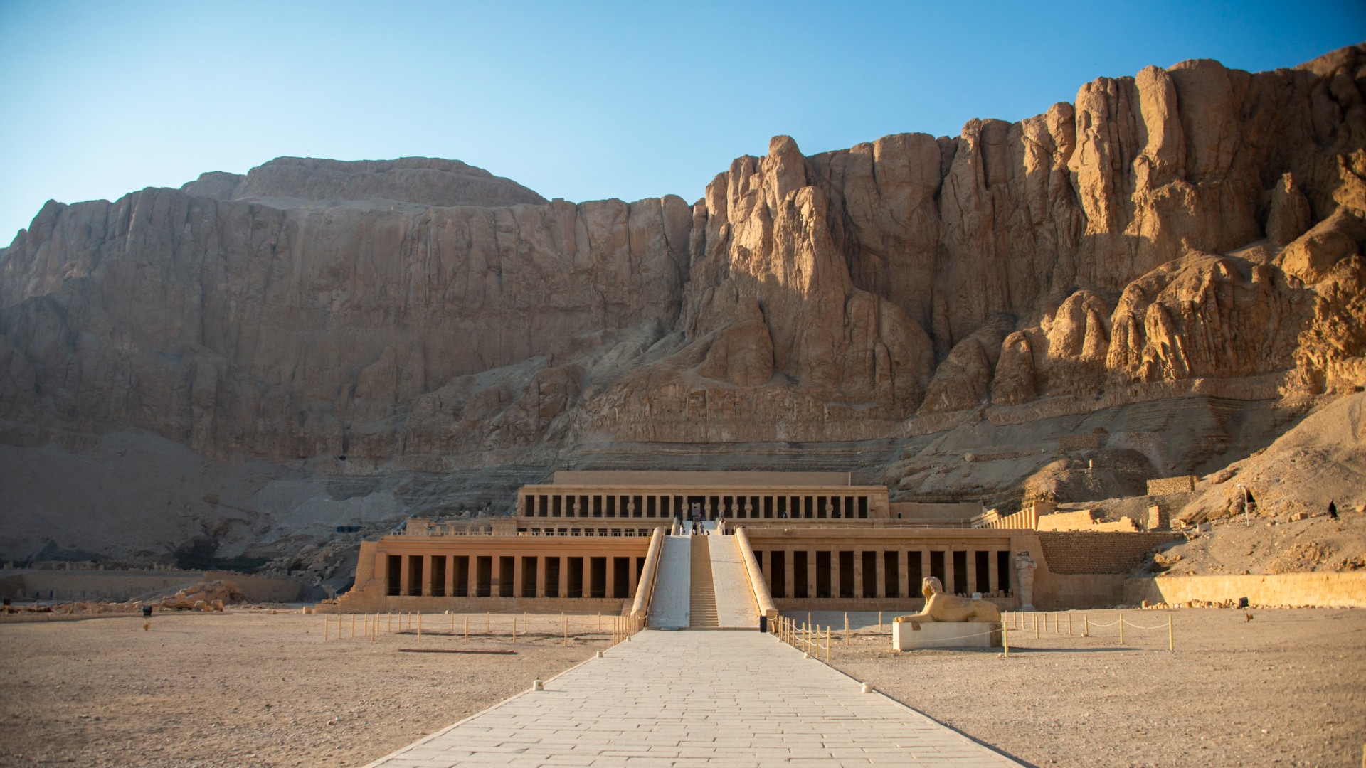 Hatshepsut&amp;rsquo;s ancient Egyptian temple stands at the end of a long stone walkway beneath towering desert cliffs, evoking a quiet, sunlit grandeur.