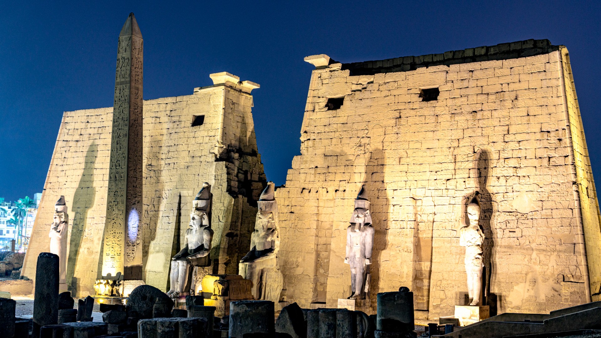 The entrance of Luxor Temple at night, featuring a tall obelisk and four large statues in front of stone pylons.