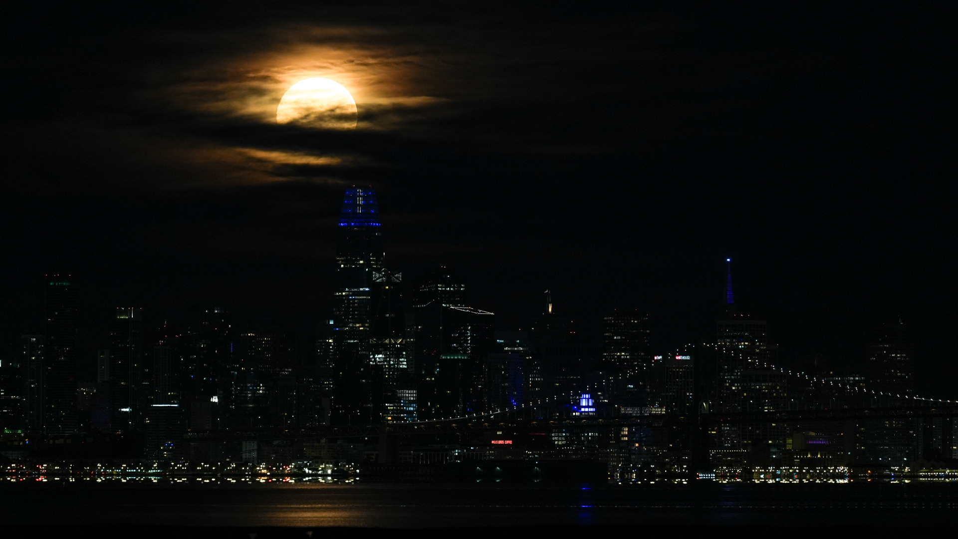 A yellow full moon glows in a cloudy sky above a city skyline lined with skyscrapers at night.