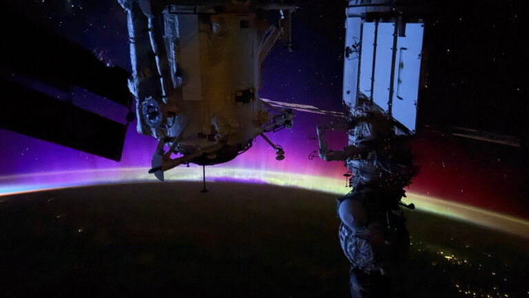 The night side of Earth is photographed from the ISS with its curved edge wreathed in colorful aurora. Solar panels and parts of the spacecraft and space station are also visible.