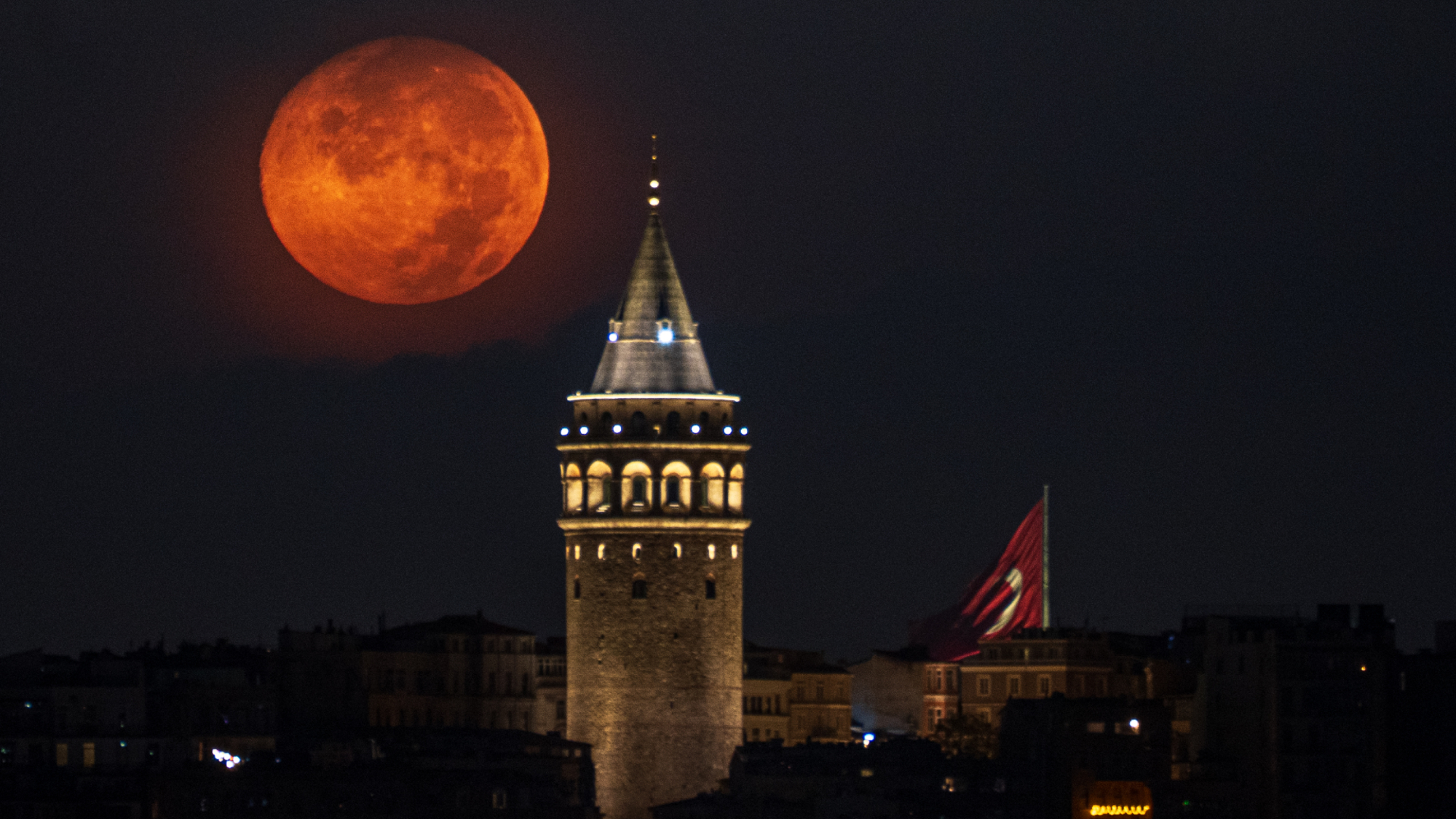 A red full moon is photographed shining next to an illuminated medieval tower at night.